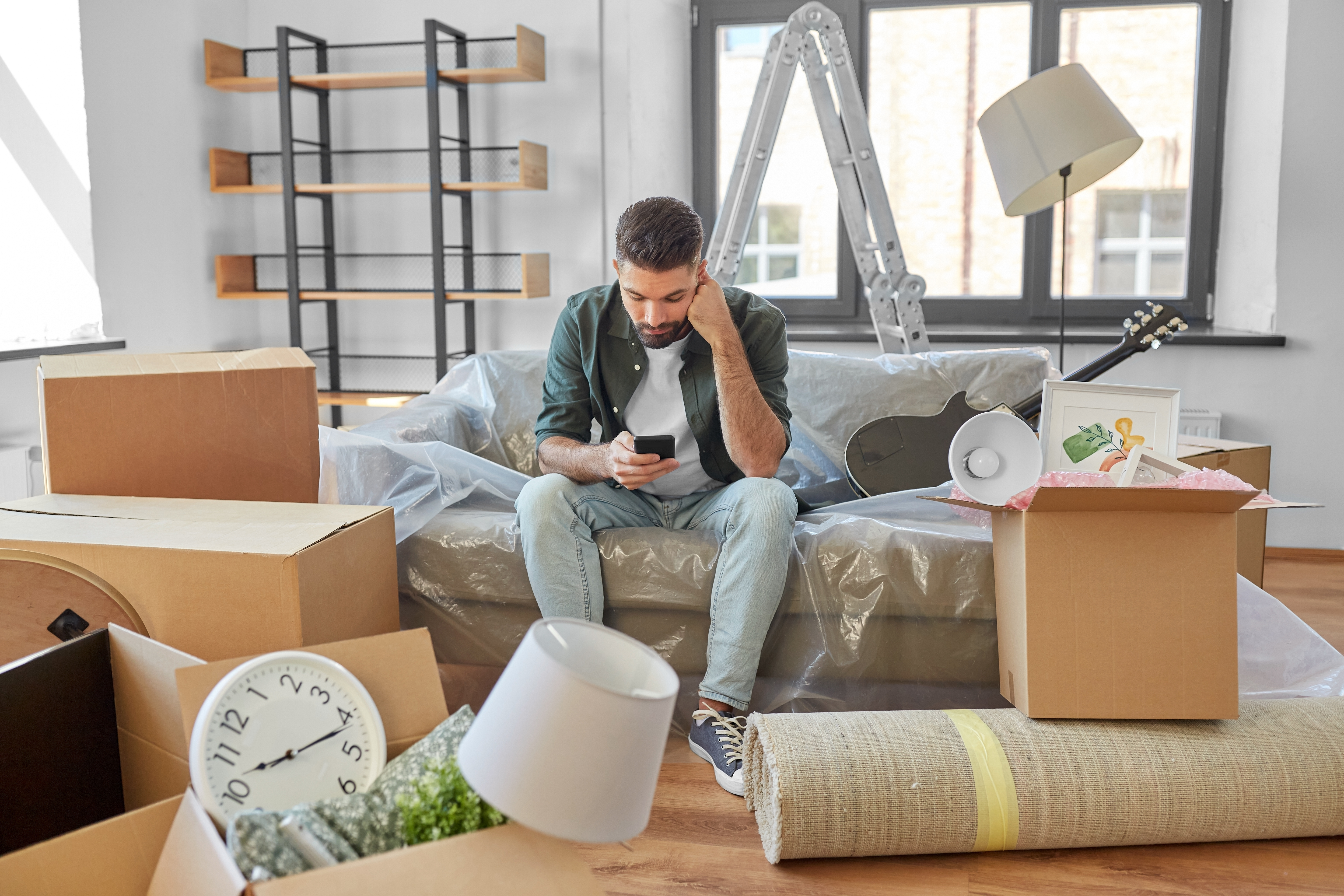 sad man with smartphone and boxes at new home